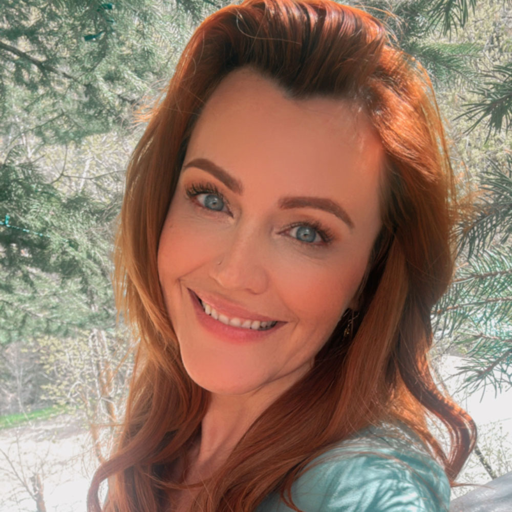 A woman with long red hair smiles at the camera while standing outdoors near green pine trees.